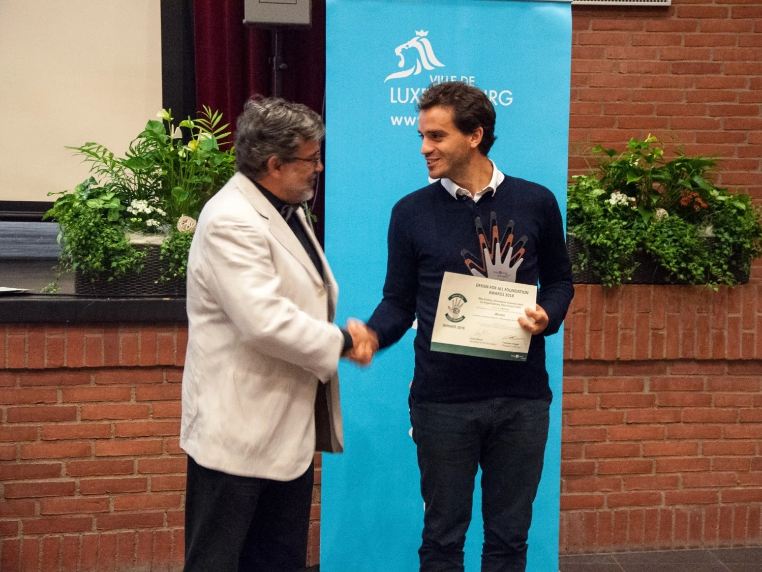 Benoît Beroud and Francesc Aragall shake hands in front of the kakemono of the city of Luxembourg